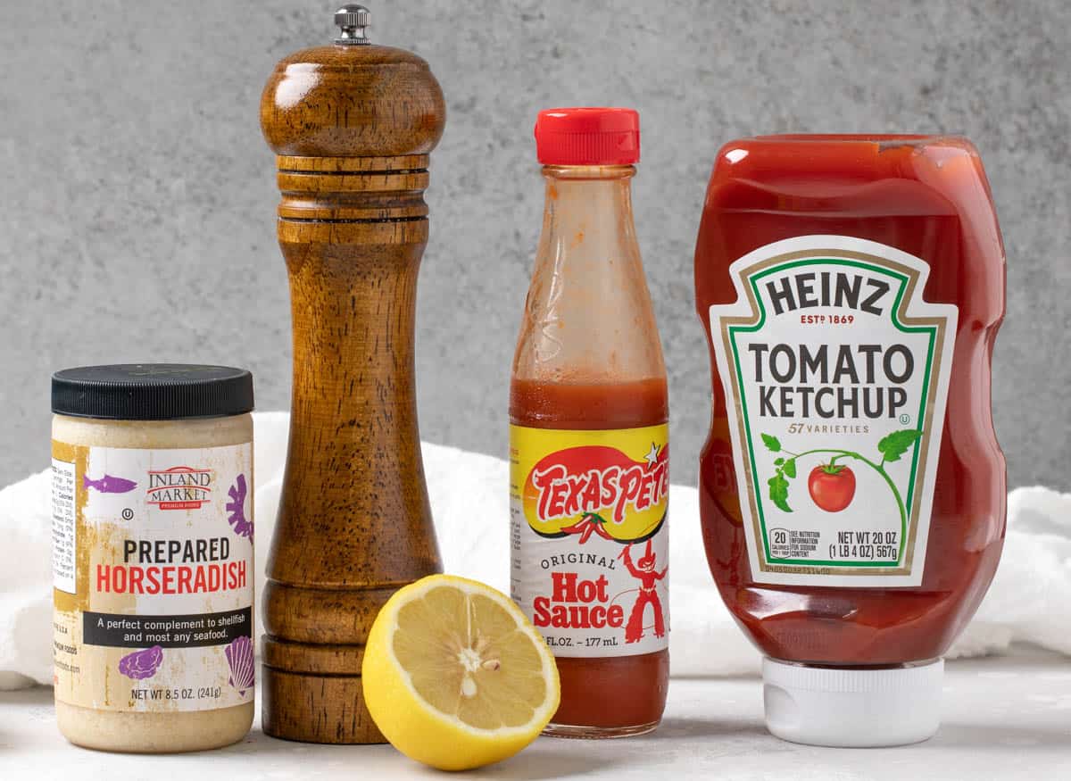 A jar of prepared horseradish, a pepper grinder, a bottle of hot sauce, a bottle of ketchup, and a lemon for making cocktail sauce.