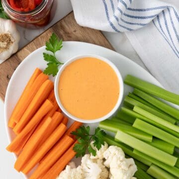 A small white bowl of red pepper aioli sauce on a platter with carrots, celery, and cauliflower.