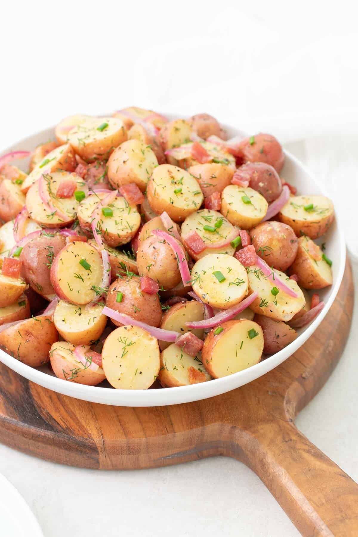 Front angled view of warm potato salad in a bowl on top of a round wooden cutting board.