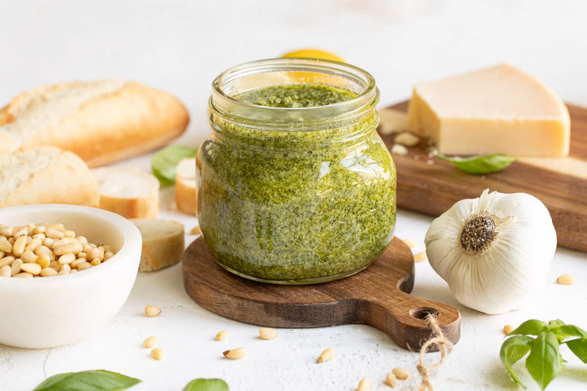 Front view of a jar of basil pesto by a garlic bulb, a jar of pine nuts, Parmesan cheese, and a French baguette.