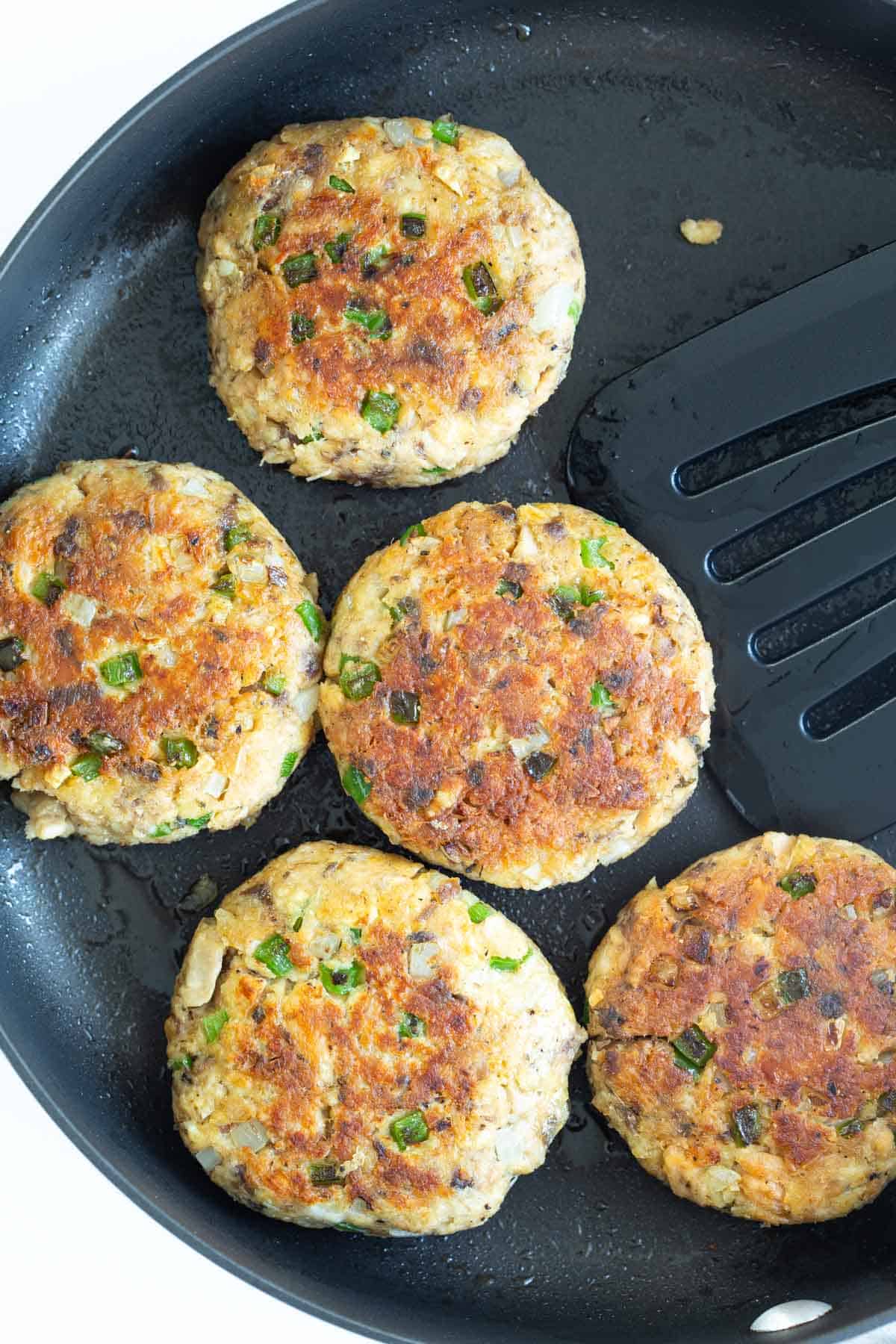 Overhead view of five salmon patties in a nonstick skillet with a spatula.