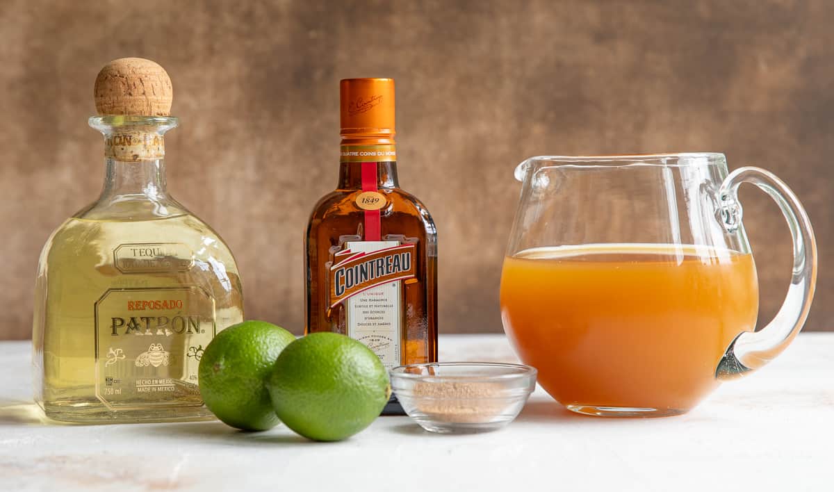 Front view of ingredients for making an apple cider margarita: tequila, limes, Cointreau, apple cider, and a cinnamon mixture for rimming the glass.