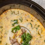 Overhead view of a ladle in an oval crock pot of Zuppa Toscana soup. Overlay text is at the top of the image.