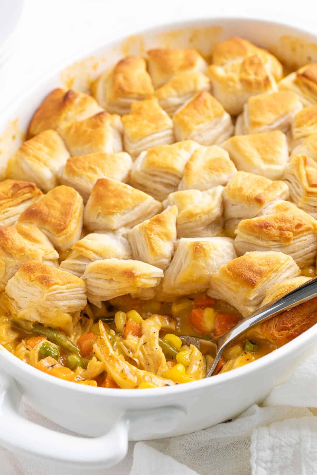 Front close-up view of a spoon in a baking dish with cheesy chicken pot pie topped with canned biscuits.