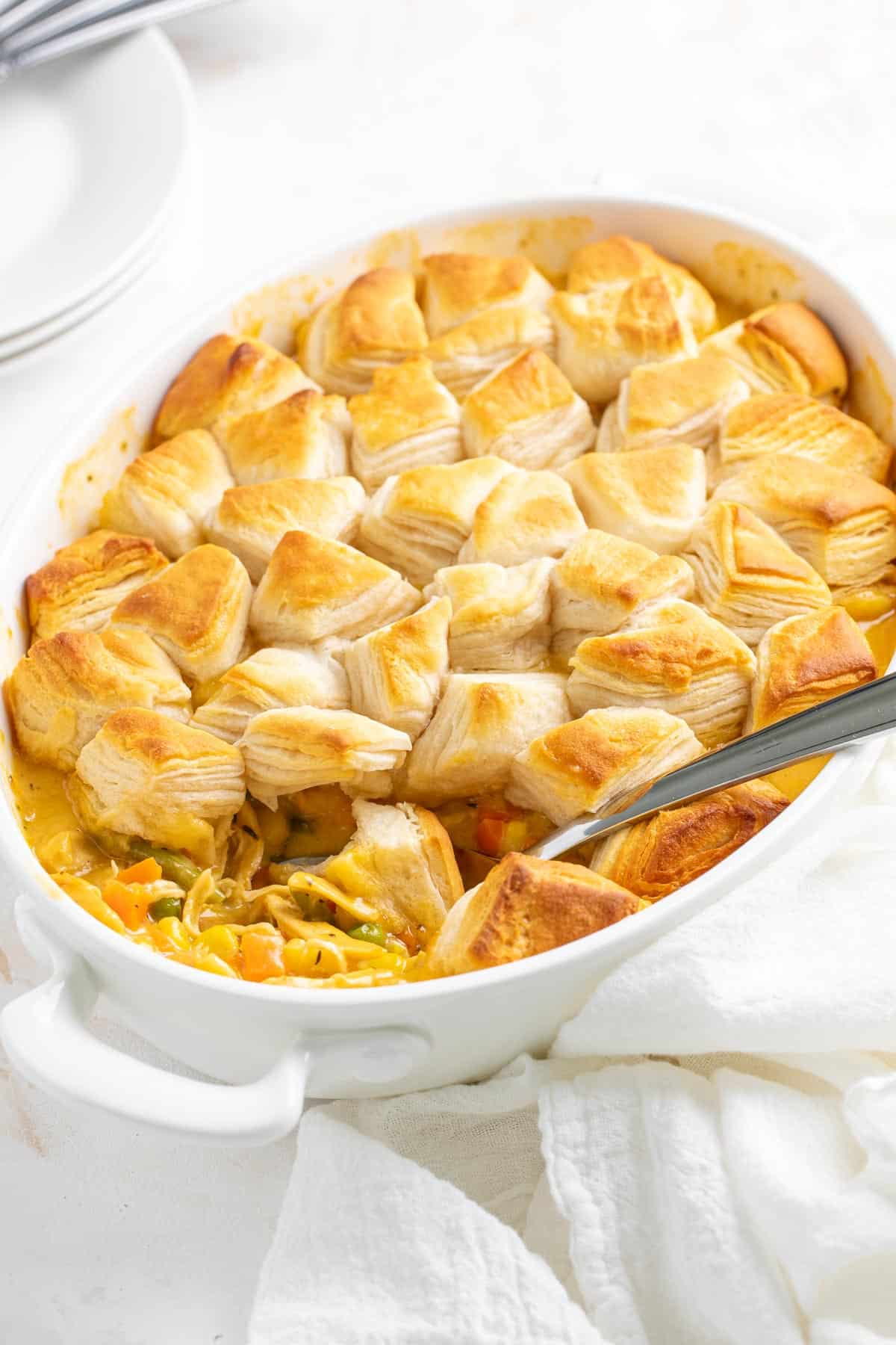 Cheesy chicken pot pie topped with quartered canned biscuits in an oval white baking dish with a serving spoon.