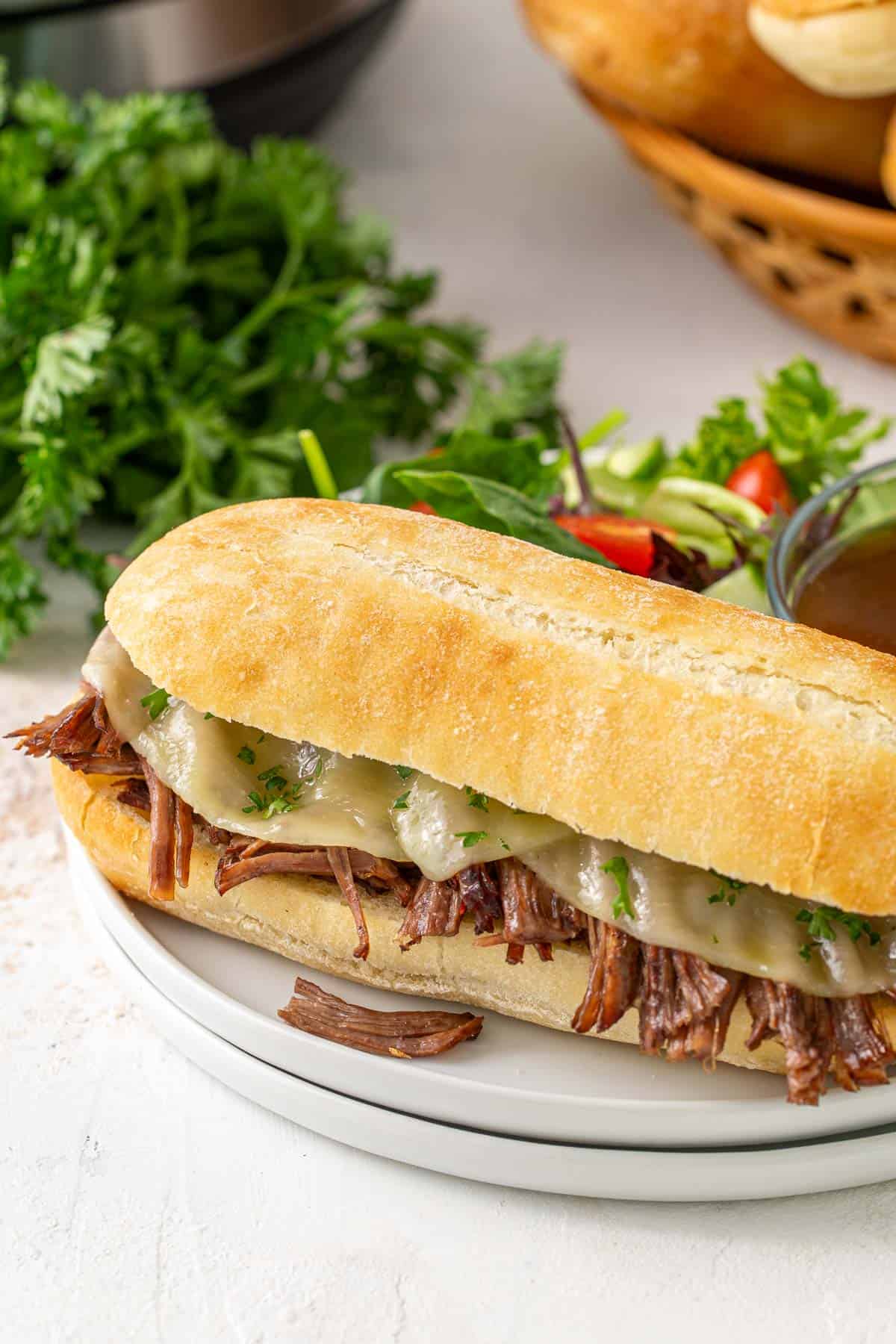 Angled view of a French dip sandwich on a white plate with a bowl of au jus and a garden salad.