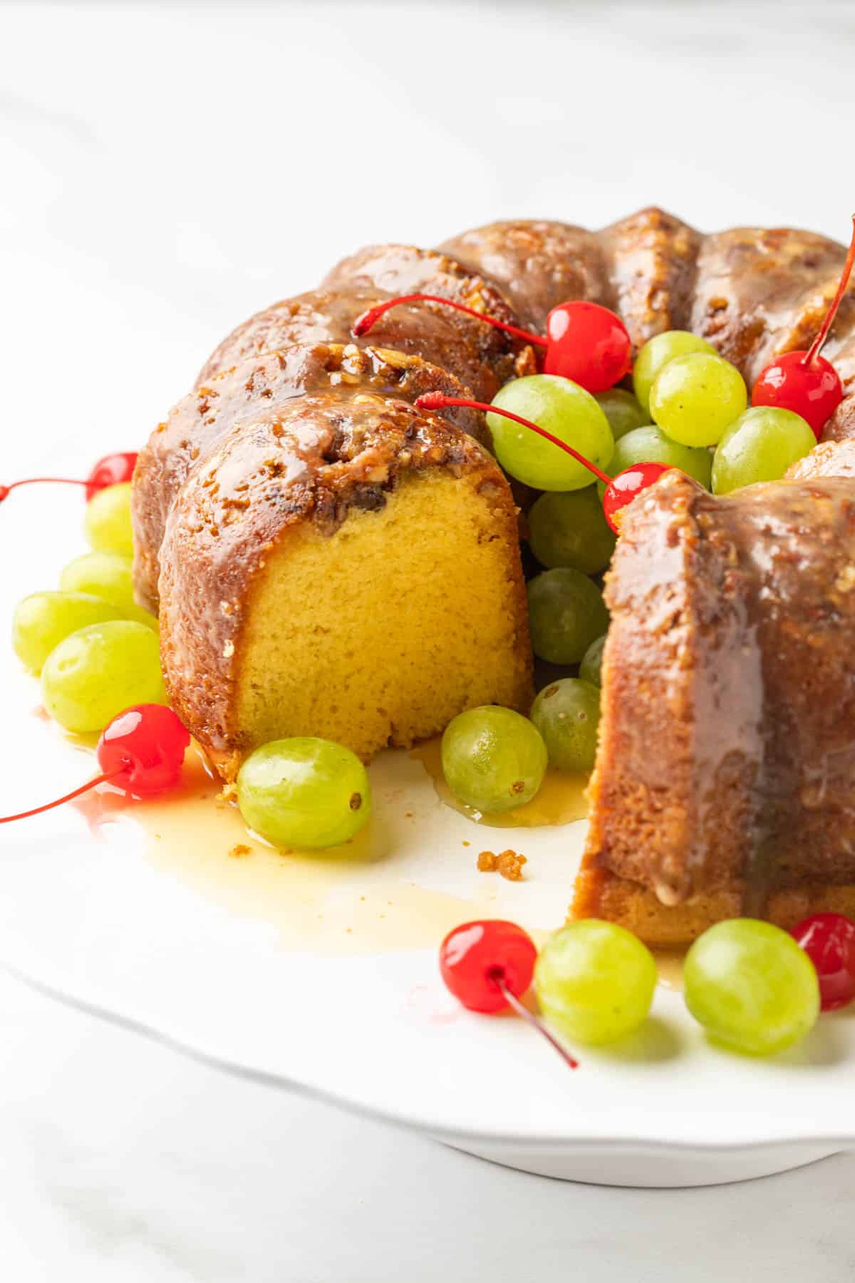 A sliced rum cake garnished with cherries and grapes on a white cake stand.