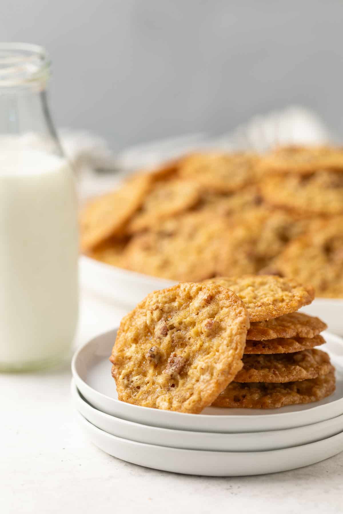 Front view of oatmeal lace cookies on a small white plate. A glass of milk is in the background.