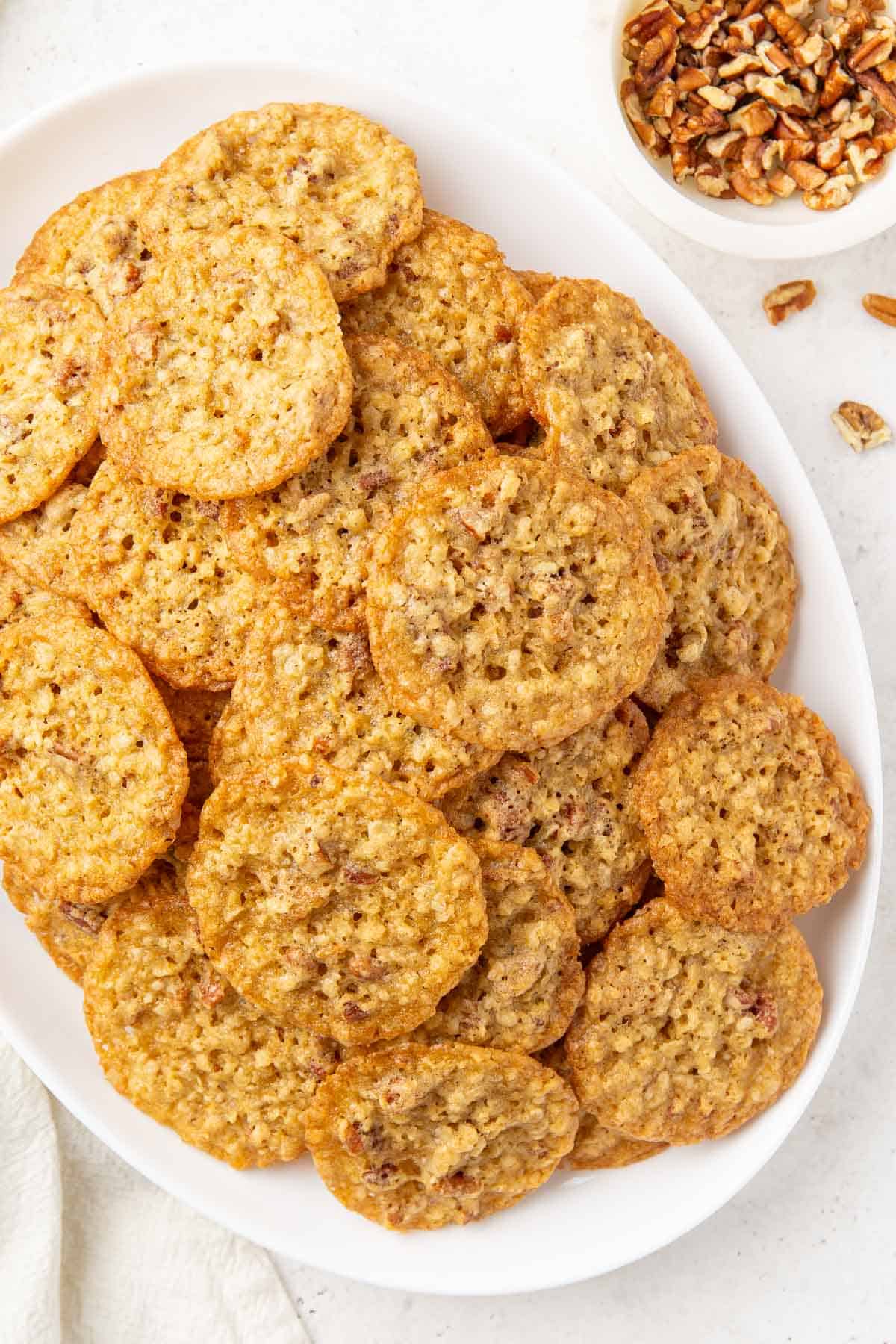 Overhead view of oatmeal lace cookies on an oval white platter beside a small bowl of chopped pecans.