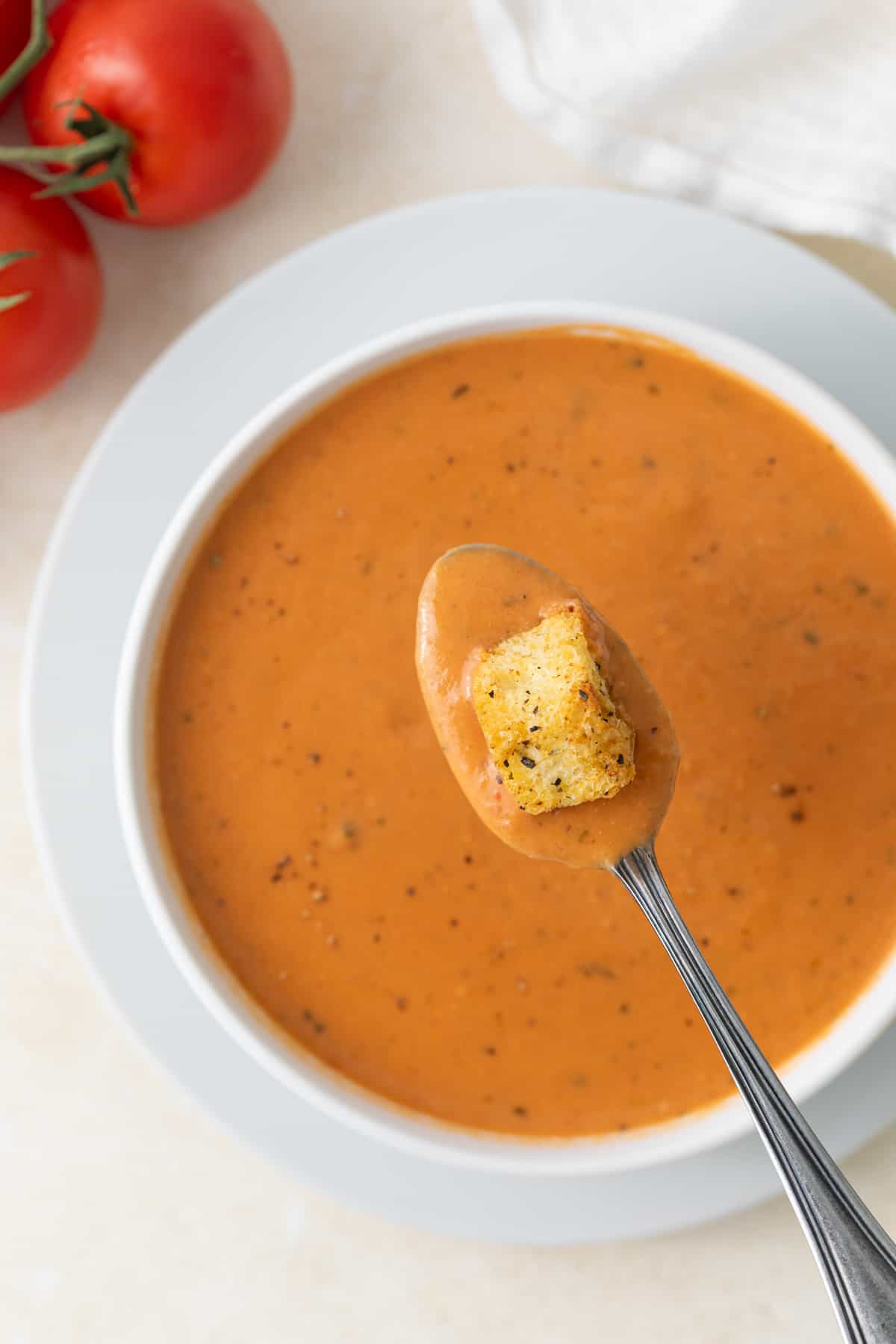 A spoonful of creamy tomato soup with a crouton over a bowl of the soup.