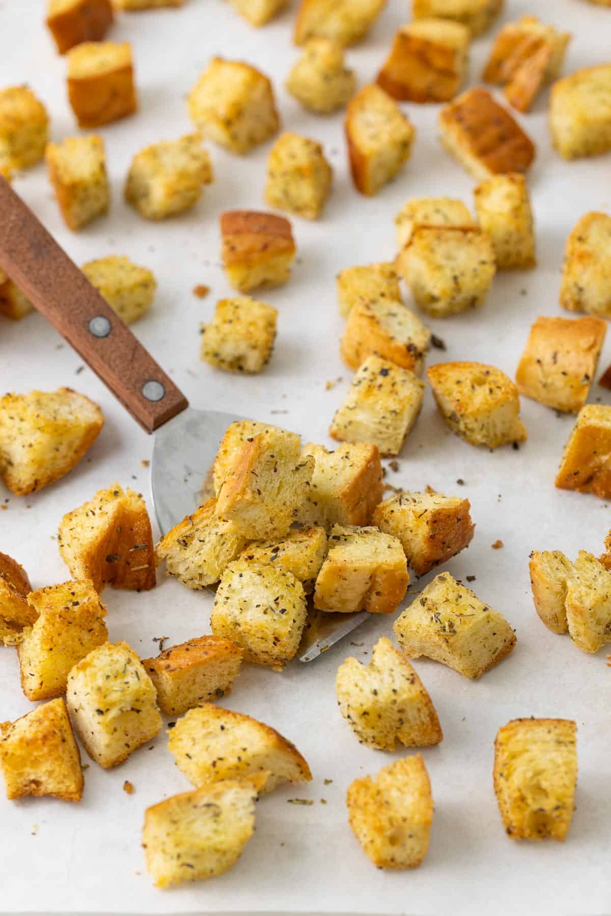 A spatula scoops up croutons on a baking sheet lined with parchment paper.