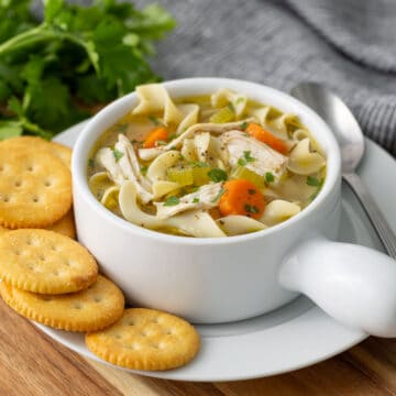 A bowl of chicken noodle soup on a plate with crackers.