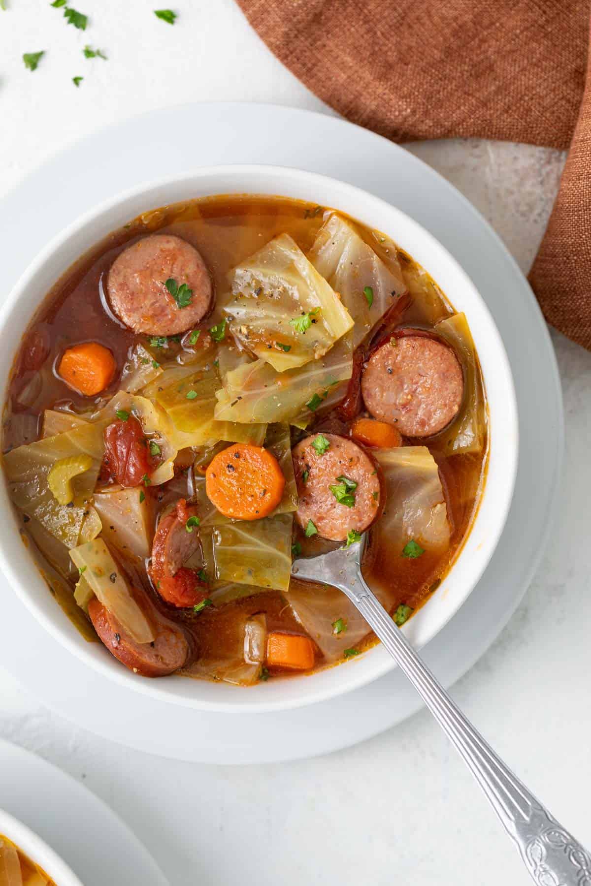 A spoon in a white bowl of cabbage soup with smoked sausage.