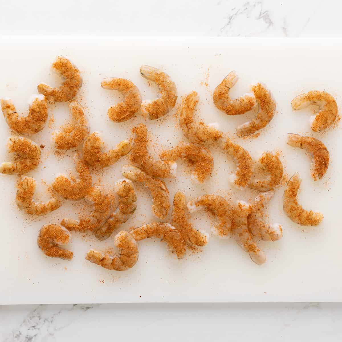 Uncooked peeled shrimp seasoned with cajun seasoning on a white cutting board.