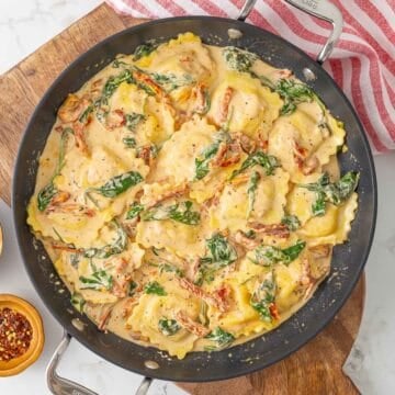 Creamy Tuscan ravioli in a skillet beside a red and white striped kitchen towel.