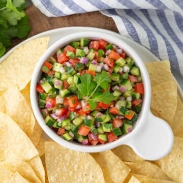 Cucumber salsa in a white bowl on a platter with tortilla chips.