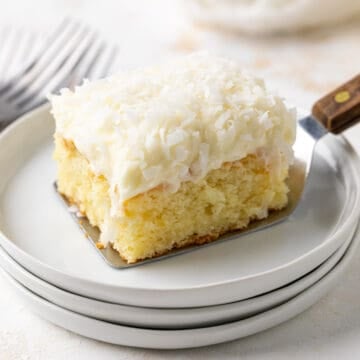 A slice of coconut sheet cake on a spatula is placed on a white dessert plate.