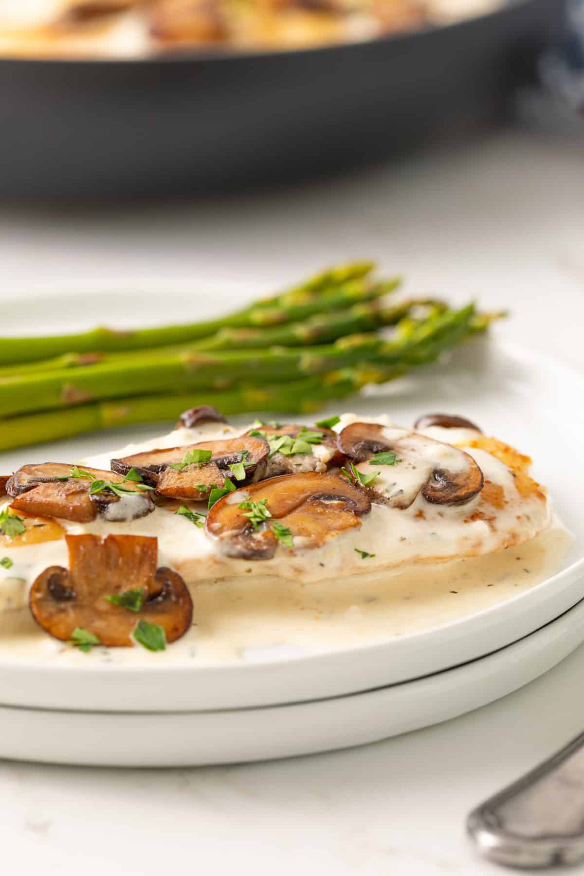 A chicken breast cutlet topped with mushroom sauce on a white plate with asparagus spears.