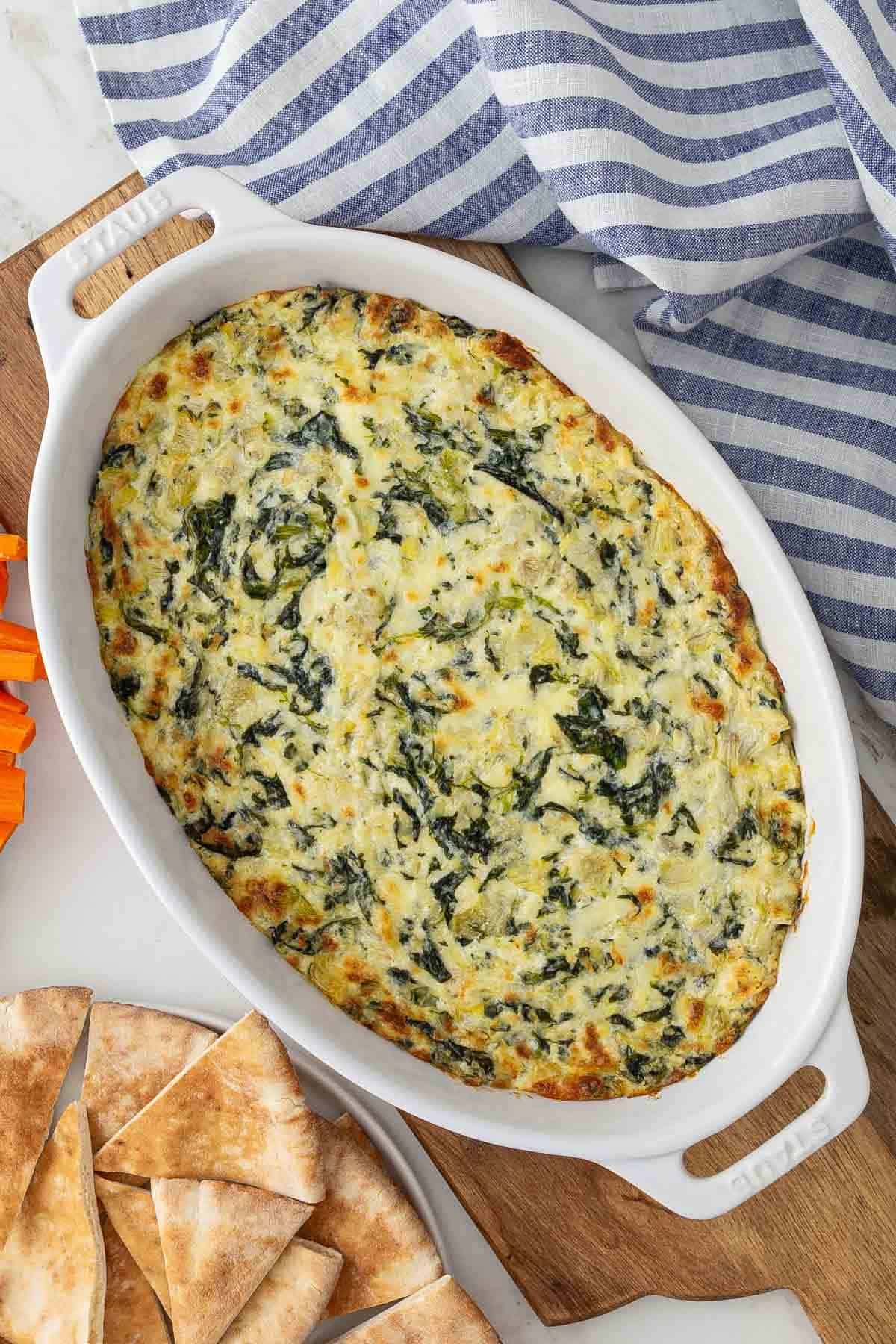 Overhead view of spinach artichoke dip in a baking dish beside plates of vegetables and bread.