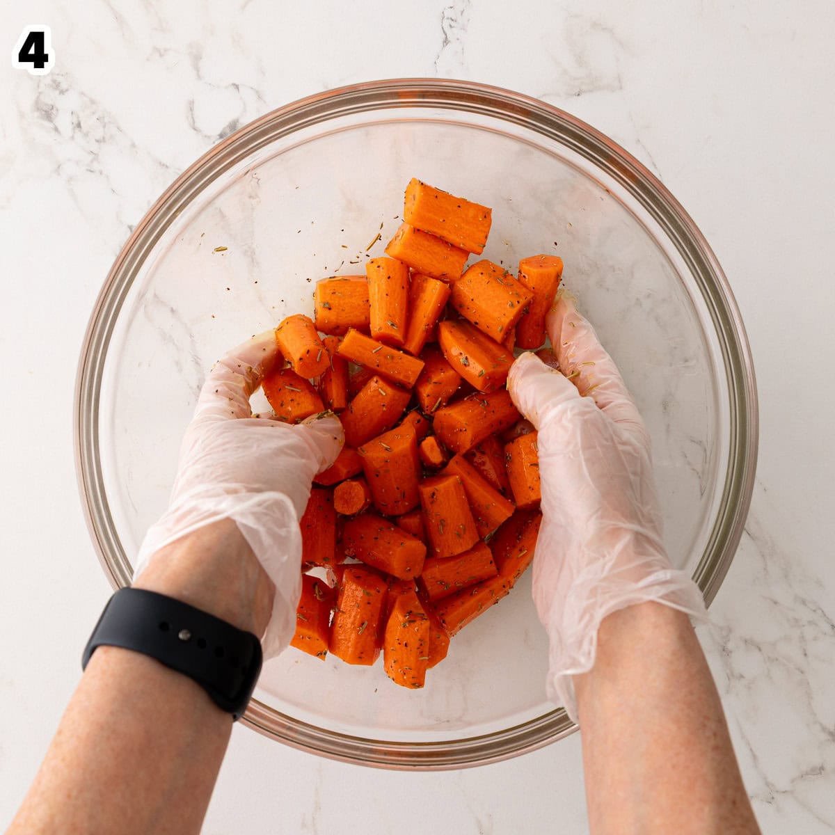Cut carrot pieces are being tossed with oil, herbs, and seasonings in a glass bowl.