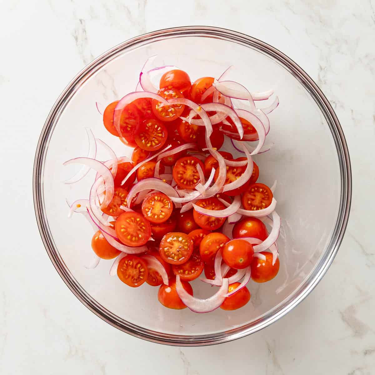 Overhead view of halved cherry tomatoes and sliced red onion in a glass bowl.
