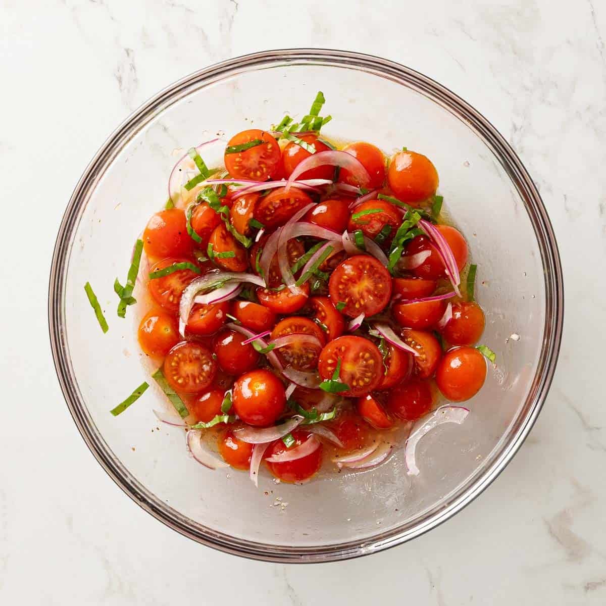 Overhead view of halved cherry tomatoes, sliced red onion, and fresh basil tossed in a dressing in a glass bowl.